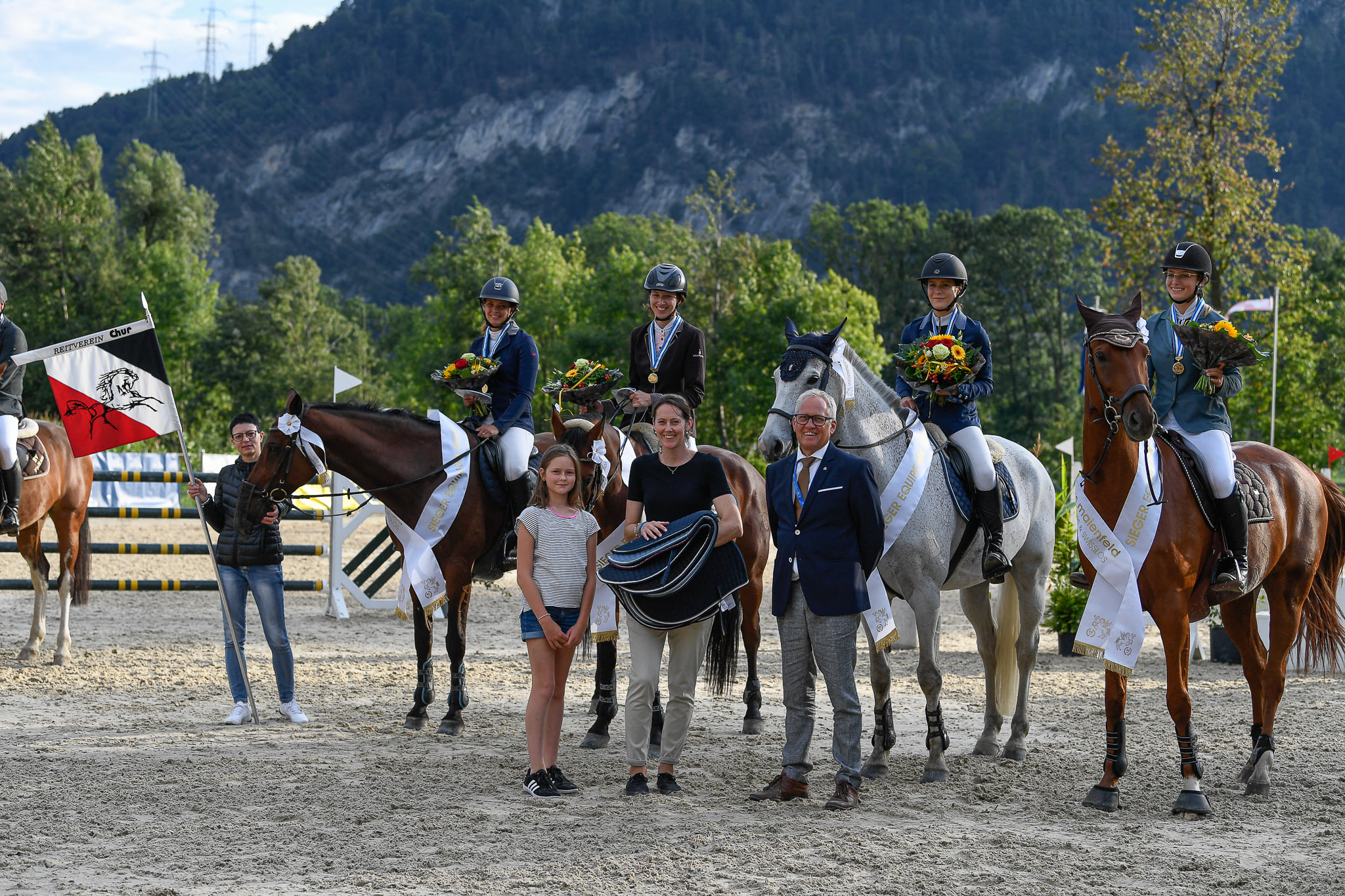 Siegerinnen eines Events auf ihren Pferden mit Blumenstrauss und Medaille, drei Personen stehen im Vordergrund davor